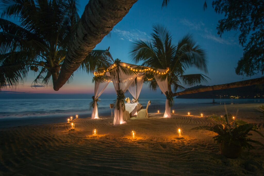 Private candlelit beachfront dining setup beneath palm trees at sunset.